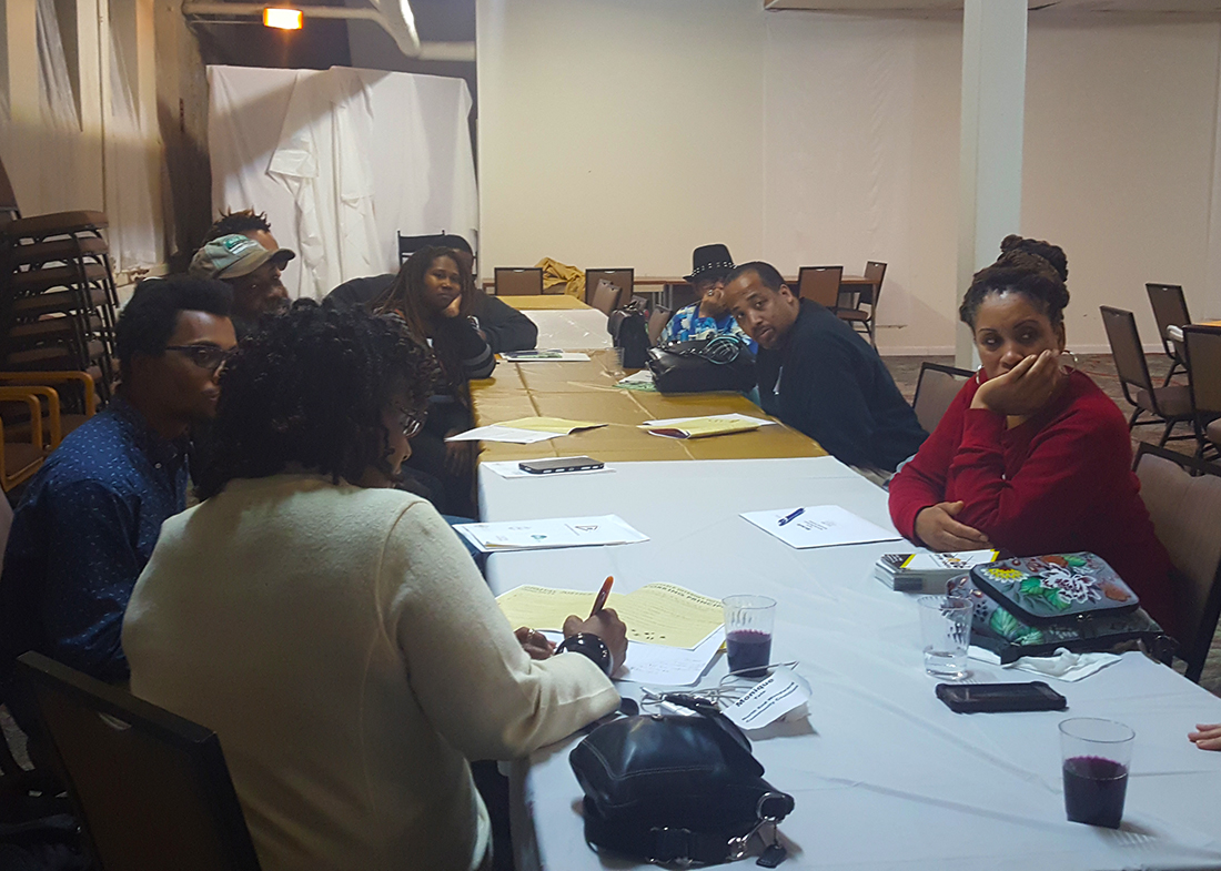 Nine community members sitting at a long table, papers sitting in front of them. Some are writing, and some are looking toward a speaker